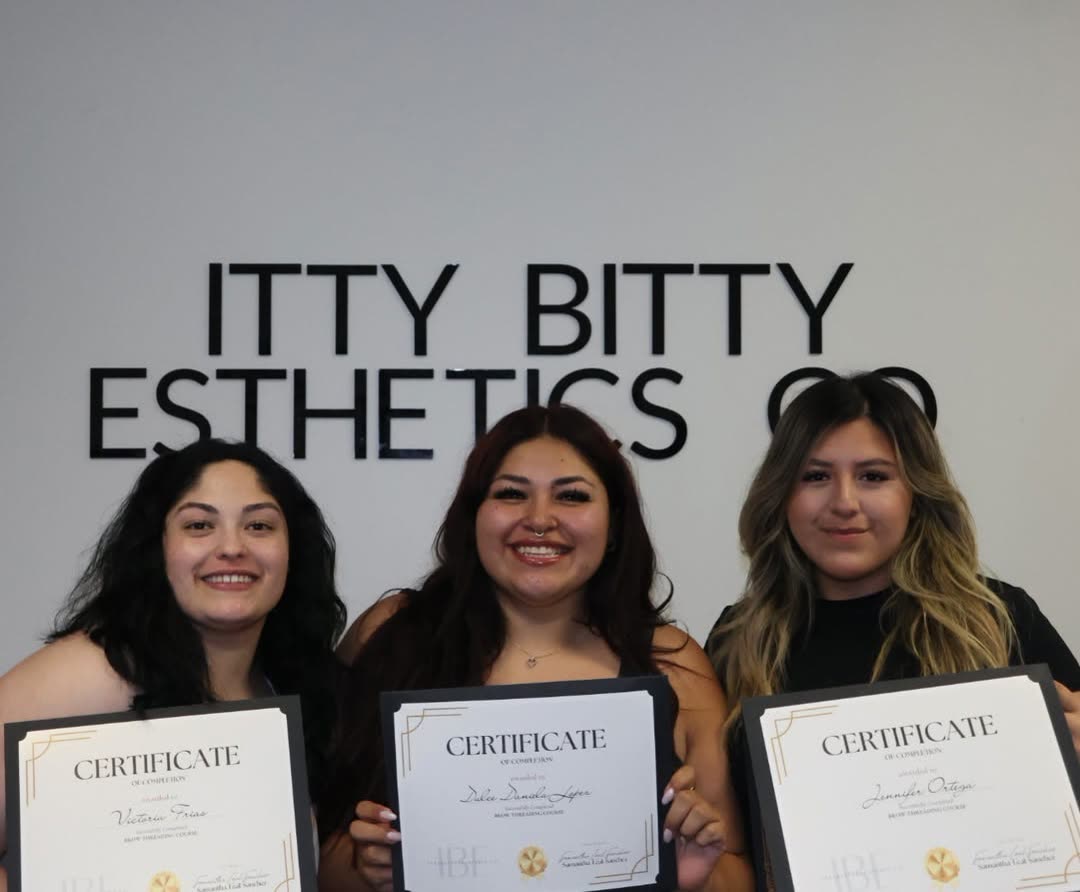 Three graduates with their certificates under the Itty Bitty Esthetics Co. sign
